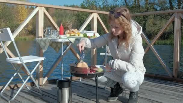 Mujer joven en ropa de abrigo, prepara verduras y carne en la parrilla, utiliza el teléfono, un perro juega cerca, un picnic en la orilla del río en un puente de madera, un fin de semana, clima frío, recreación al aire libre — Vídeos de Stock