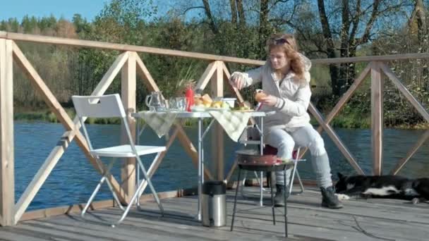 Mujer joven en ropa de abrigo, preparando verduras y carne en la parrilla, preparando una hamburguesa, un perro jugando cerca, un picnic en la orilla del río en un puente de madera, un fin de semana, clima frío, al aire libre — Vídeos de Stock