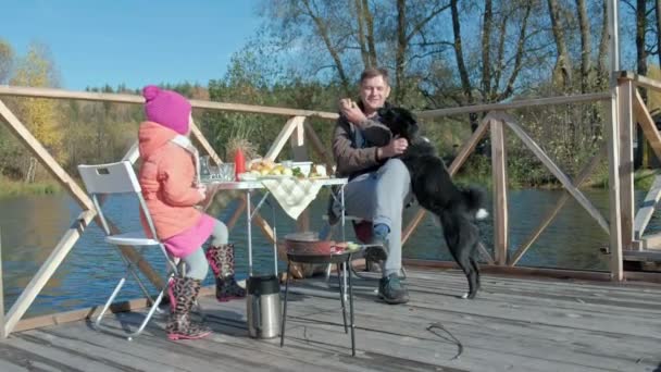 Père et fille, homme mûr et petite fille, en vêtements chauds, assis à la table, manger, cuire des légumes sur le gril, près d'un chien affamé, un pique-nique sur la rive d'une rivière sur un pont en bois, week-end, froid — Video