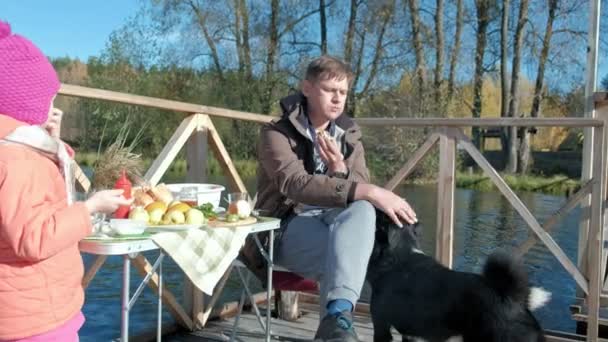Батько і дочка, зрілий чоловік і маленька дівчинка, сидять за столом, їдять, готують овочі на грилі, біля голодного собаки, пікнік на березі річки на дерев'яному мосту, вихідні, холодні — стокове відео