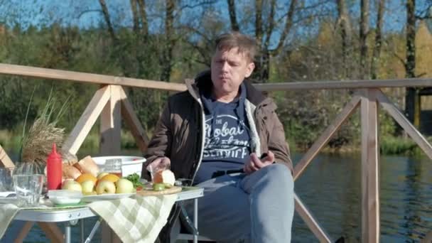 Reifer Mann in warmer Kleidung, am Tisch sitzend, Burger essen, neben einem hungrigen Hund, Picknick am Flussufer auf einer Holzbrücke, Wochenende, kaltes Wetter, Zelten, Tourismus — Stockvideo