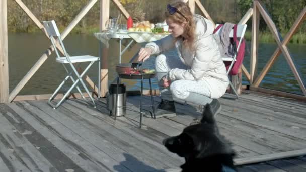 Молодая женщина в теплой одежде, готовит овощи и мясо на гриле, готовит бургер, поблизости играет собака, пикник на берегу реки на деревянном мосту, выходные, холодная погода, на открытом воздухе — стоковое видео