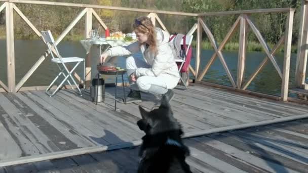 Junge Frau in warmer Kleidung, Gemüse und Fleisch auf dem Grill zubereiten, Burger zubereiten, Hund in der Nähe spielen, Picknick am Flussufer auf einer Holzbrücke, ein Wochenende, kaltes Wetter, draußen — Stockvideo