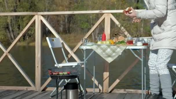 Mujer joven en ropa de abrigo, preparando verduras y carne en la parrilla, preparando una hamburguesa, un perro jugando cerca, un picnic en la orilla del río en un puente de madera, un fin de semana, clima frío, al aire libre — Vídeos de Stock