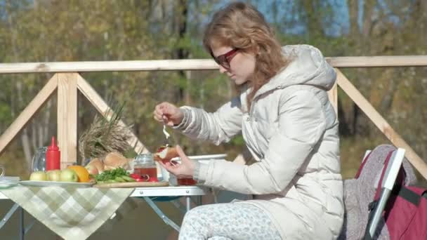 Jeune femme en vêtements chauds, préparant légumes et viande sur le gril, préparant un hamburger, un chien jouant à proximité, un pique-nique sur la rive d'une rivière sur un pont en bois, un week-end, temps froid, extérieur — Video