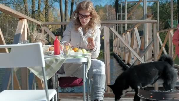 Jonge vrouw in warme kleren, eten van een hamburger, een hond speelt in de buurt, een picknick aan de rivier op een houten brug, weekend, koud weer, camping, toerisme — Stockvideo