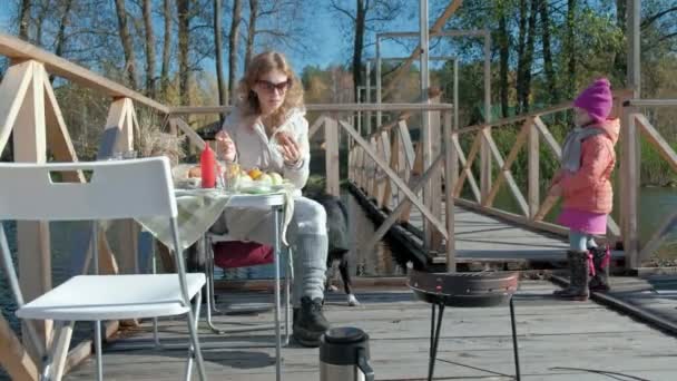 Mamma och dotter, ung kvinna i varma kläder äta en hamburgare, nära det finns en liten flicka med en hund, en picknick vid floden banken på en träbro, helg, kallt väder, friluftsliv, turism — Stockvideo