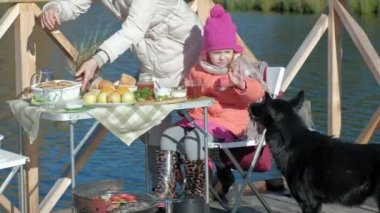 Anne ve kızı, Genç bir kadın ve küçük bir kız sıcak giysiler içinde bir kutu açılır, bir köpek yakın, nehrin kenarında piknik çalış ahşap köprü, hafta sonları, soğuk hava, kamp, turizm üzerinde