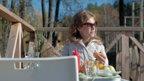 Ung kvinna i varma kläder, äta en hamburgare, en hund spelar i närheten, en picknick vid floden på en träbro, helg, kallt väder, camping, turism — Stockvideo