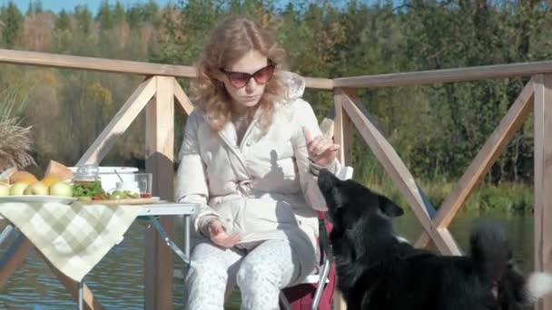 Nuori nainen lämpimissä vaatteissa, syö hampurilaisen, koira leikkii lähellä, piknik joen rannalla puisella sillalla, viikonloppuna, kylmä sää, retkeily, matkailu — kuvapankkivideo