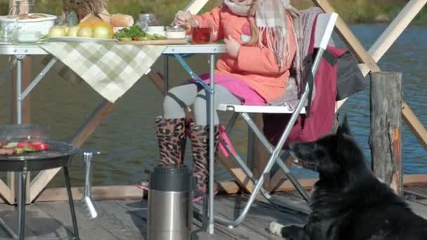 Девочка в теплой одежде, ест блины, пьет чай, играет собака поблизости, пикник у реки на деревянном мосту, выходные, холодная погода, кемпинг, туризм — стоковое видео