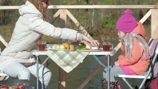 Μητέρα και κόρη, νεαρή γυναίκα και το μικρό κορίτσι σε ζεστά ρούχα, κάθεται στο τραπέζι, πίνοντας τσάι και τρώγοντας, πικ-νικ από τον ποταμό σε μια ξύλινη γέφυρα, Σαββατοκύριακο, κρύο, κάμπινγκ, τουρισμός — Αρχείο Βίντεο