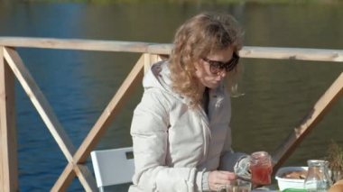 masada oturan, çay içme, reçel, bir ahşap köprü, hafta sonu, soğuk hava, kamp, turizm nehir kıyısında piknik ile pancakes yemek sıcak giysiler içinde genç kadın