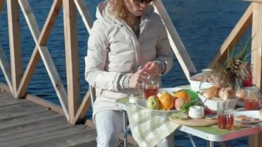 masada oturan, çay içme, reçel, bir ahşap köprü, hafta sonu, soğuk hava, kamp, turizm nehir kıyısında piknik ile pancakes yemek sıcak giysiler içinde genç kadın