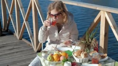 masada oturan, çay içme, reçel, bir ahşap köprü, hafta sonu, soğuk hava, kamp, turizm nehir kıyısında piknik ile pancakes yemek sıcak giysiler içinde genç kadın