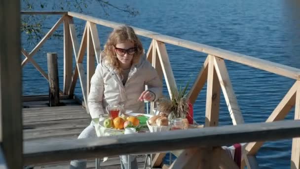mladá žena v teplé oblečení, sedící u stolu, pití čaje, jíst palačinky s marmeládou, piknik u řeky na dřevěný most, víkend, chladné počasí, camping, Turistika
