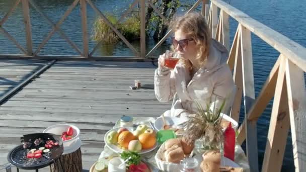 Νεαρή γυναίκα σε ζεστά ρούχα, κάθεται στο τραπέζι, πίνοντας τσάι και τρώγοντας τηγανίτες με μαρμελάδα, πικ-νικ από τον ποταμό σε μια ξύλινη γέφυρα, Σαββατοκύριακο, κρύο, κάμπινγκ, τουρισμός — Αρχείο Βίντεο