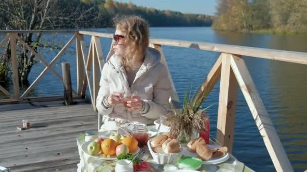 Mujer joven en gafas de sol calientes, sentado a la mesa, beber té, comer panqueques con mermelada, picnic junto al río en un puente de madera, fin de semana, clima frío, camping, turismo — Vídeos de Stock