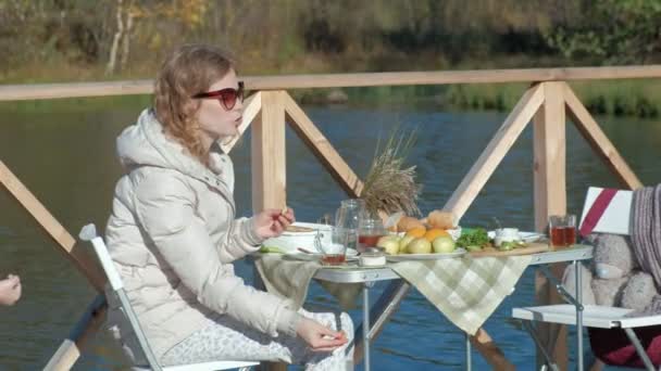 Giovane donna in occhiali da sole, in abiti caldi, seduta a un tavolo, bere tè, mangiare frittelle con marmellata, nutrire un cane, pic-nic sul fiume su un ponte di legno, fine settimana, freddo, campeggio, turismo — Video Stock