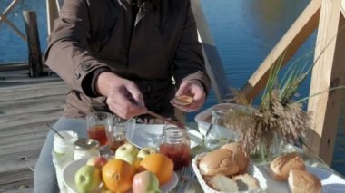 Olgun adam koyar reçel krep, yiyor, bir ahşap köprü, hafta sonu, soğuk hava, kamp, turizm nehir kıyısında piknik sıcak giysiler içinde