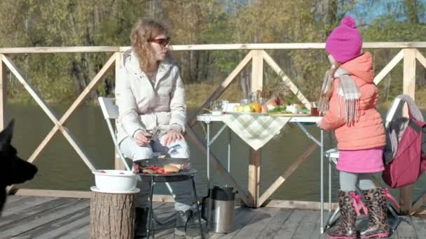 Madre e hija, mujer joven en gafas de sol, ropa de abrigo, sentarse a la mesa, beber té, comer panqueques con mermelada, al lado de la chica que juega con un perro, picnic junto al río en un puente de madera — Vídeos de Stock