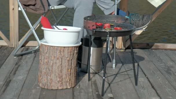 Reifer Mann in warmer Kleidung, am Tisch sitzend, Hamburger essen, Picknick am Flussufer auf einer Holzbrücke, Wochenende, kaltes Wetter, Erholung im Freien, Tourismus — Stockvideo