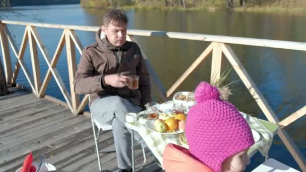 Uomo maturo, in abiti caldi, seduto a tavola, mangiare un hamburger, l'aggiunta di ketchup, pic-nic sulla riva del fiume su un ponte di legno, fine settimana, freddo, campeggio, turismo — Video Stock