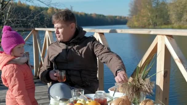 Padre e hija, hombre y chica maduros, en ropa de abrigo, sentado a la mesa, alimentando a la chica con panqueques, picnic en la orilla del río en un puente de madera, fin de semana, clima frío, recreación al aire libre — Vídeo de stock