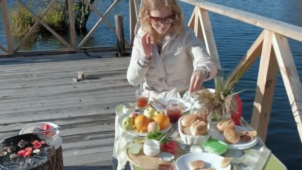 Mujer joven en gafas de sol calientes, sentado a la mesa, beber té, comer panqueques con mermelada, picnic junto al río en un puente de madera, fin de semana, clima frío, camping, turismo — Vídeo de stock