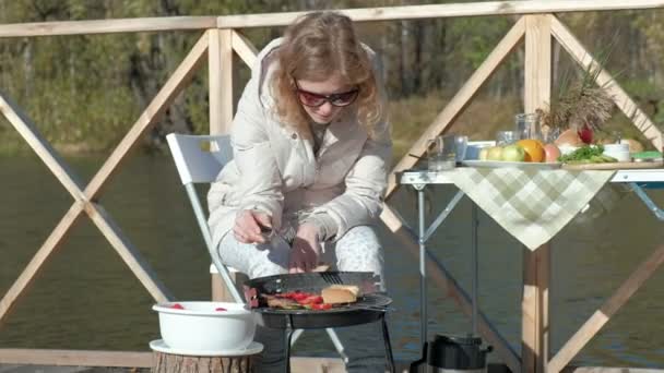 Молодая женщина в теплой одежде, готовит овощи и мясо на гриле, готовит бургер, поблизости играет собака, пикник на берегу реки на деревянном мосту, выходные, холодная погода, на открытом воздухе — стоковое видео