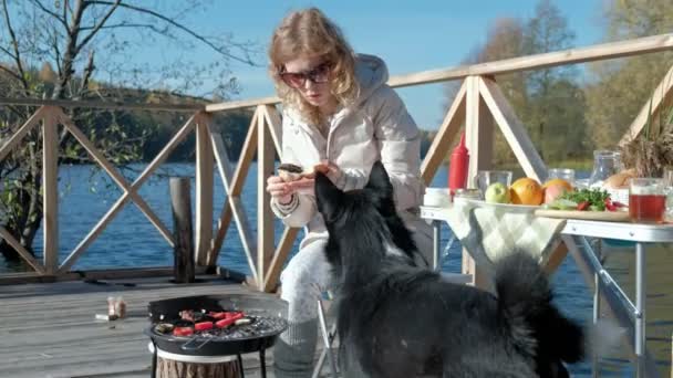 Junge Frau in warmer Kleidung, Gemüse und Fleisch auf dem Grill, Hamburger kochen, neben einem hungrigen Hund, Picknick am Flussufer auf einer Holzbrücke, Wochenende, kaltes Wetter, Erholung im Freien — Stockvideo