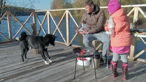 Vader en dochter, volwassen man en meisje, in warme kleren, zitten aan de tafel, voeden van de hond, picknick op de rivier bank op een houten brug, weekend, koud weer, outdoor recreatie, toerisme — Stockvideo