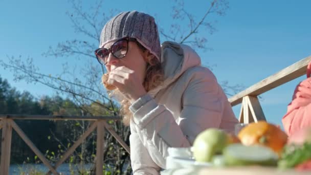 Ung kvinna i varma kläder, äta en hamburgare, en hund spelar i närheten, en picknick vid floden på en träbro, helg, kallt väder, camping, turism — Stockvideo