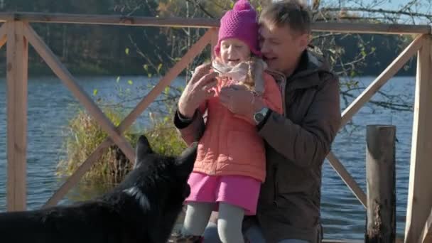 Тато і дочка, зрілий чоловік і маленька дівчинка, в теплому одязі, посмішка, годувати собаку, пікнік на березі річки на дерев'яному мосту, вихідні, холодна погода, кемпінг, туризм, щастя — стокове відео