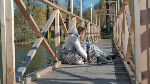 秋の川の近くの橋の上の女性は、黒犬を近くに歩いている本を読んでいます。 — ストック動画
