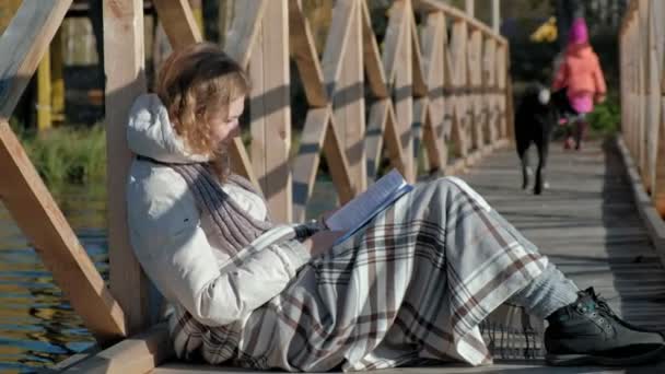 Uma mulher na ponte perto do rio de outono está lendo um livro um cão preto e uma menina estão andando nas proximidades — Vídeo de Stock