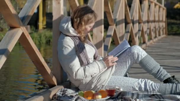 Una mujer en el puente cerca del río de otoño lee un libro y bebe té — Vídeos de Stock