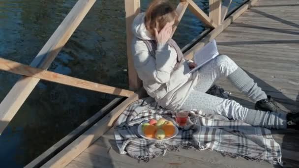 Una mujer en el puente cerca del río de otoño lee un libro y bebe té — Vídeo de stock