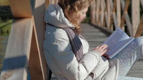 Una mujer en el puente cerca del río de otoño lee un libro y bebe té — Vídeo de stock