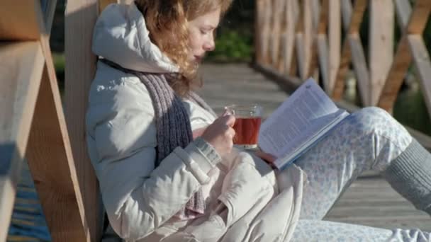 Una mujer en el puente cerca del río de otoño lee un libro y bebe té — Vídeos de Stock