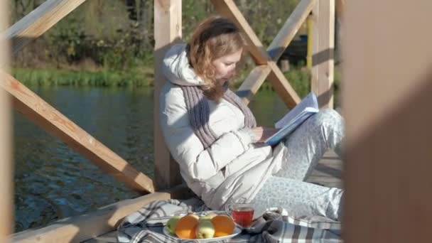 Eine Frau auf der Brücke am Herbstfluss liest ein Buch und trinkt Tee — Stockvideo