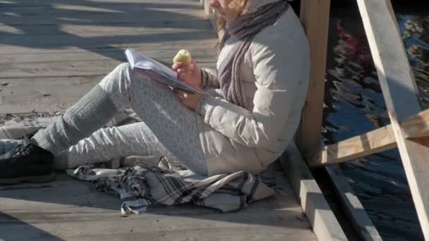 Mulher na ponte perto do rio outono lê um livro e come uma maçã — Vídeo de Stock