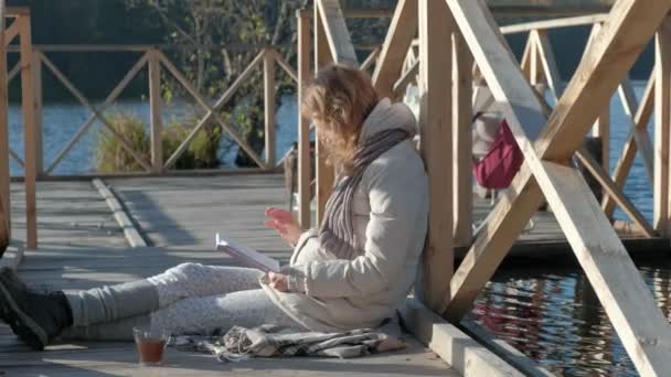 Femme sur le pont près de la rivière d'automne lit un livre et mange une pomme — Video