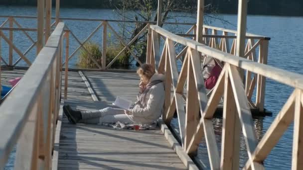 Vrouw op de brug in de buurt van de rivier in de herfst een boek leest en eet een appel — Stockvideo