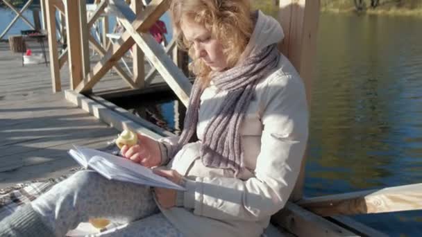 Mujer en el puente cerca del río de otoño lee un libro y se come una manzana — Vídeos de Stock