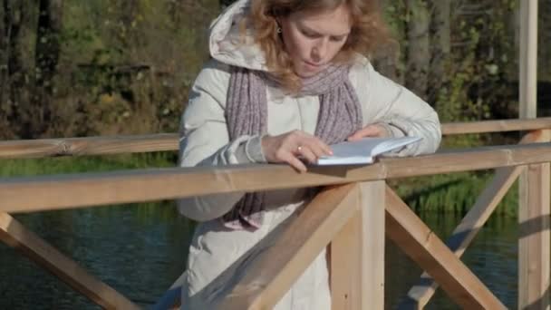 Γυναίκας στη γέφυρα κοντά στον ποταμό φθινόπωρο διαβάζει ένα βιβλίο — Αρχείο Βίντεο