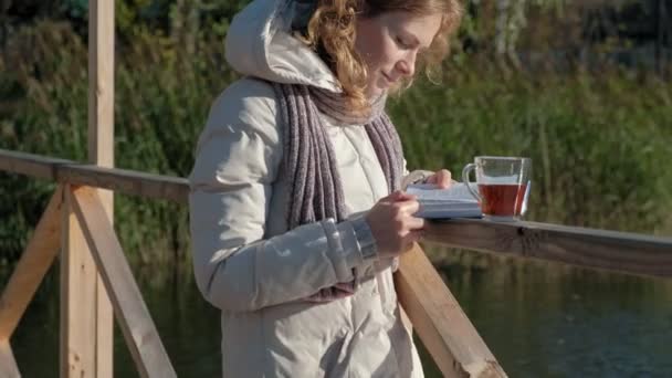 Une femme sur le pont près de la rivière d'automne lit un livre et boit du thé — Video