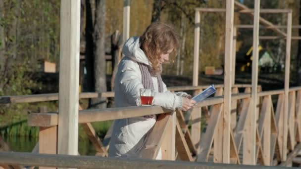 En kvinna på bron nära floden hösten läser en bok och dricker te — Stockvideo