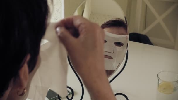 Стара жінка дивиться на зморшки у дзеркалі. Вона відкриває косметичну маску і кладе її на обличчя. Догляд за обличчям. Пластична хірургія та ін'єкції колагену. Реалістичні образи своїми руками — стокове відео