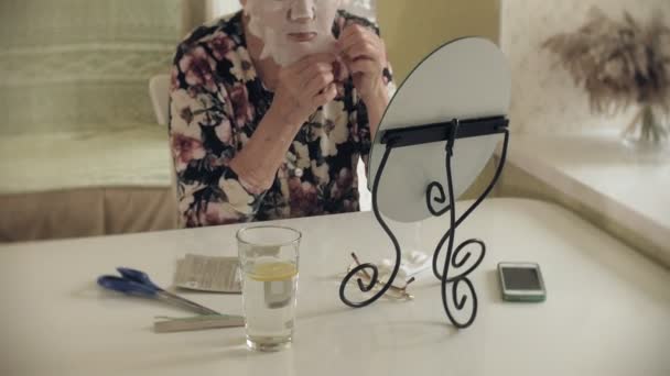 Yaşlı bir kadının aynaya bakar ve onun yüzüne bir kozmetik kumaş maskesi uygular. Dış cephe bakım. Plastik cerrahi ve Kollajen enjeksiyonları. Kendi kusurları ile gerçekçi görüntüler. — Stok video